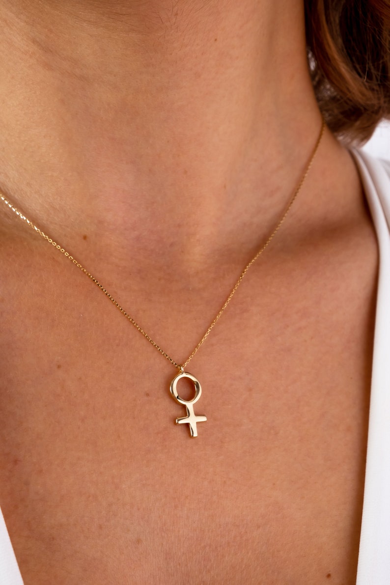 K&ouml;nnte beinhalten: Eine goldene Halskette mit einem schlichten, minimalistischen Anh&auml;nger in Form des weiblichen Symbols.
