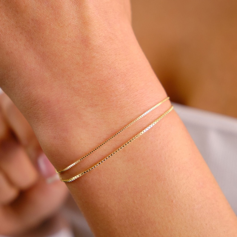 May include: Two gold chain bracelets layered on a wrist. The bracelets are made of a delicate box chain.