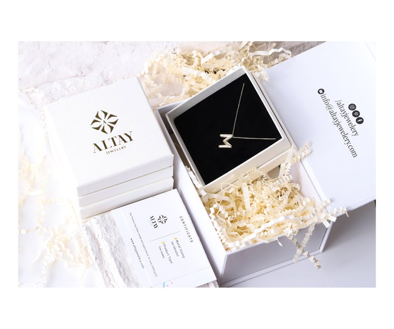 Op de afbeelding: Een witte doos met een gouden logo met de tekst "Altay Jewelry" is open om een witte doos te onthullen met een gouden ketting met een letter "M" hanger. De ketting ligt op een zwart fluwelen kussen. De doos is omgeven door wit kreukelpapier.