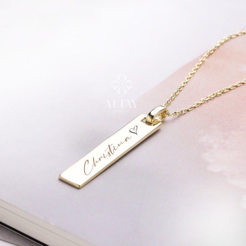 May include: Gold necklace with a rectangular pendant engraved with the name "Christina" and a small heart symbol.
