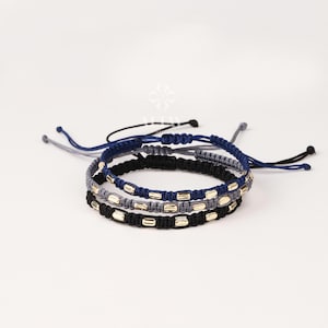 May include: Three macrame bracelets in navy blue, black, and gray, each adorned with gold-colored rectangular beads. The bracelets are knotted and adjustable, with black and gray cords extending from the ends. The bracelets are stacked on top of each other.