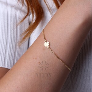 Puede incluir: Una delicada pulsera dorada con un dije de tr&eacute;bol de cuatro hojas. La pulsera est&aacute; sujeta a una fina cadena y se lleva en el brazo. El dije de tr&eacute;bol est&aacute; centrado en el brazo. El fondo es una camisa blanca.