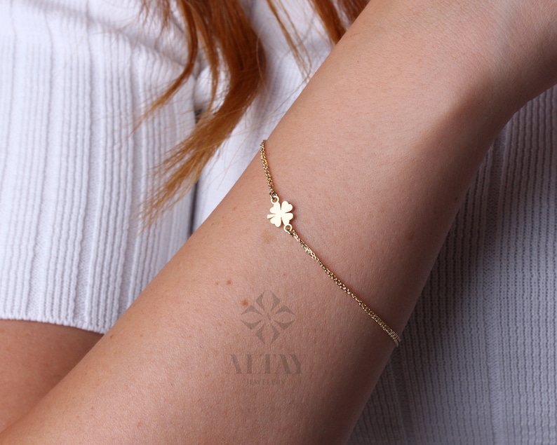 Puede incluir: Una delicada pulsera dorada con un dije de tr&eacute;bol de cuatro hojas. La pulsera est&aacute; sujeta a una fina cadena y se lleva en el brazo. El dije de tr&eacute;bol est&aacute; centrado en el brazo. El fondo es una camisa blanca.