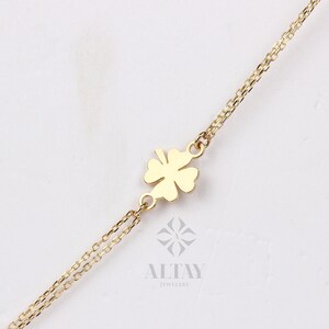 Puede incluir: Un collar dorado con un colgante de tr&eacute;bol de cuatro hojas. El tr&eacute;bol est&aacute; centrado en una delicada cadena. El collar se muestra sobre un fondo blanco. Se ve el nombre de la marca "ALTAY JEWELRY".