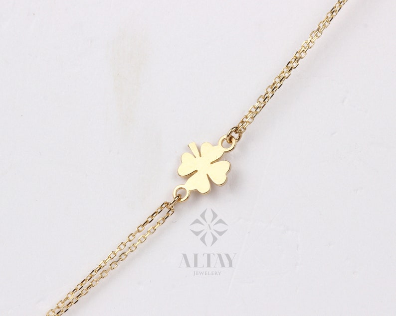 Puede incluir: Un collar dorado con un colgante de tr&eacute;bol de cuatro hojas. El tr&eacute;bol est&aacute; centrado en una delicada cadena. El collar se muestra sobre un fondo blanco. Se ve el nombre de la marca "ALTAY JEWELRY".