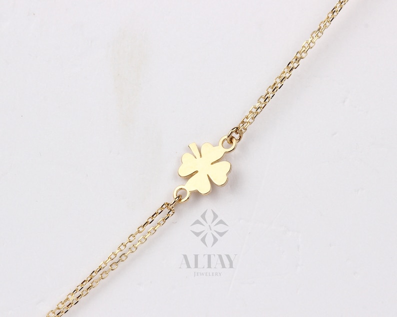 Puede incluir: Un collar dorado con un colgante de tr&eacute;bol de cuatro hojas. El tr&eacute;bol est&aacute; centrado en una delicada cadena. El collar se muestra sobre un fondo blanco. Se ve el nombre de la marca "ALTAY JEWELRY".