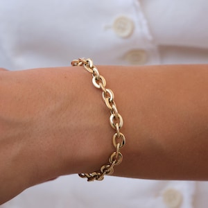 Könnte beinhalten: Ein goldfarbenes Kettengliederarmband. Das Armband besteht aus oval geformten Gliedern und wird am Handgelenk getragen. Der Hintergrund ist ein weißes Hemd. Die Glieder sind etwa 1,5 cm lang.