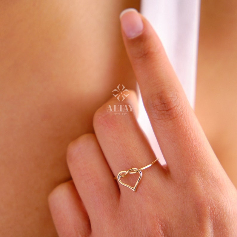 May include: A gold heart-shaped ring with a knot design. The ring is worn on a finger.