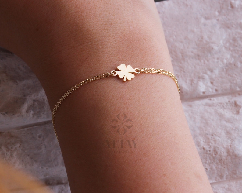 Puede incluir: Una pulsera dorada con un dije de tr&eacute;bol de cuatro hojas. La delicada pulsera de cadena se muestra en un brazo. El dije de tr&eacute;bol es el centro de atenci&oacute;n. Una joya sencilla y elegante.