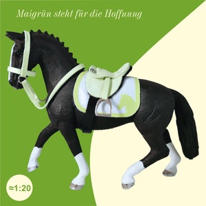 Ein schwarzes Schleich Pferd mit einemhellgrünen Sattel und einer Trense. Darüber steht der Schriftzug: Maigrün steht für die Hoffnung.