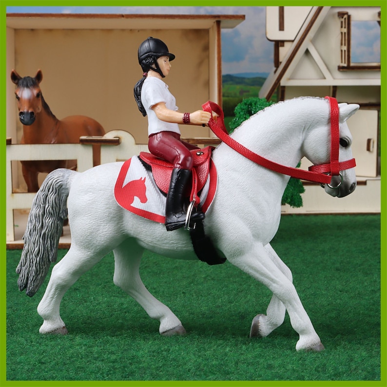 Eine Schleich Reiterin reitet ein weißes Schleich Pferd das mit einem roten Sattel gesattelt ist. Im Hintergrund ist ein Offenstall mit einem Pferd zu sehen.