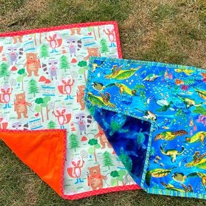May include: Two colorful baby blankets. One features a woodland scene with bears and raccoons, the other depicts sea turtles and fish. Both have a patterned design and a contrasting orange or blue backing.