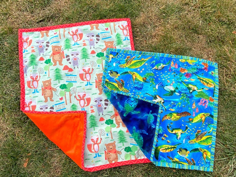 May include: Two colorful baby blankets. One features a woodland scene with bears and raccoons, the other depicts sea turtles and fish. Both have a patterned design and a contrasting orange or blue backing.