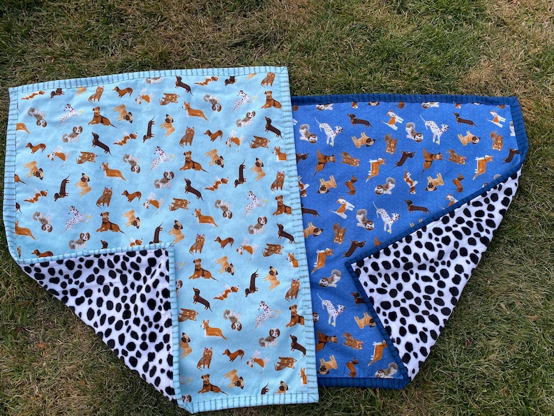 May include: Two dog-themed blankets. One is light blue with various dog breeds, the other is blue with similar dog illustrations. Both have a black and white dalmatian-spotted underside. The blankets are rectangular with a stitched border.