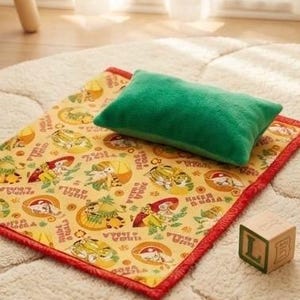 May include: A green rectangular pillow rests on a yellow mat with a red border. The mat features a repeating pattern of cartoon characters and text. A wooden block with the letter "L" is also present.