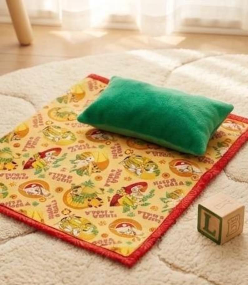 May include: A green rectangular pillow rests on a yellow mat with a red border. The mat features a repeating pattern of cartoon characters and text. A wooden block with the letter "L" is also present.