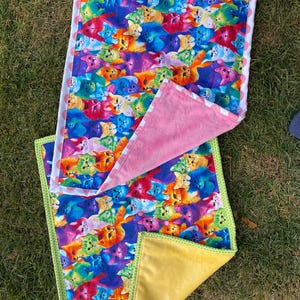 May include: Two colorful cat-themed blankets. One blanket features a vibrant pattern of cats in various colors, with a white border and pink polka dots. The other blanket has a yellow backing and green trim.