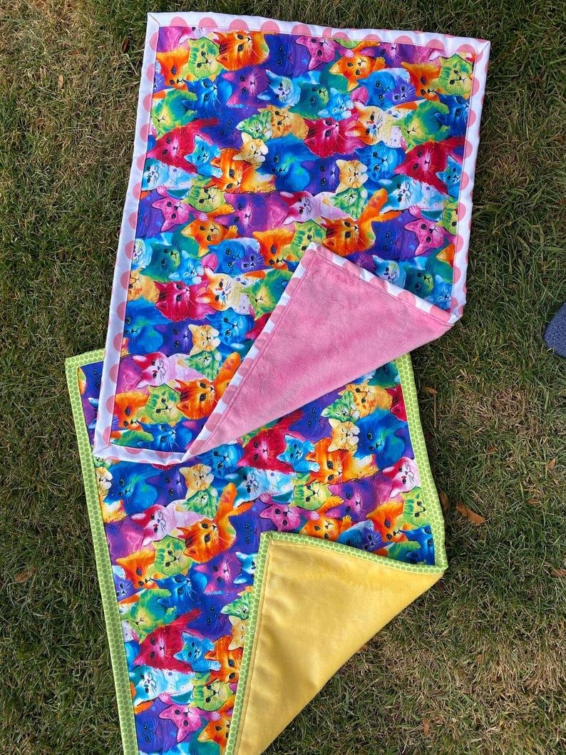 May include: Two colorful cat-themed blankets. One blanket features a vibrant pattern of cats in various colors, with a white border and pink polka dots. The other blanket has a yellow backing and green trim.