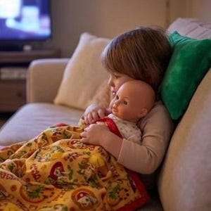 May include: A child cuddling a doll while sitting on a beige sofa. The doll is wrapped in a yellow blanket with a red border and a pattern of cartoon characters. A green pillow is behind the child.