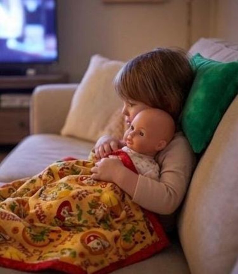 May include: A child cuddling a doll while sitting on a beige sofa. The doll is wrapped in a yellow blanket with a red border and a pattern of cartoon characters. A green pillow is behind the child.