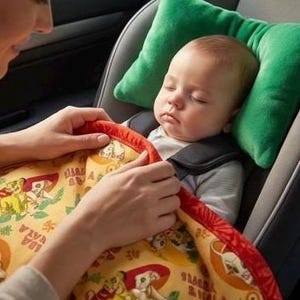 May include: A baby sleeping in a car seat, secured with a harness. The baby is nestled against a green pillow and covered with a yellow blanket featuring a cartoon design. A person's hands are gently adjusting the blanket.