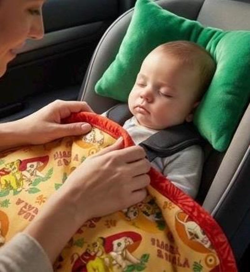 May include: A baby sleeping in a car seat, secured with a harness. The baby is nestled against a green pillow and covered with a yellow blanket featuring a cartoon design. A person's hands are gently adjusting the blanket.