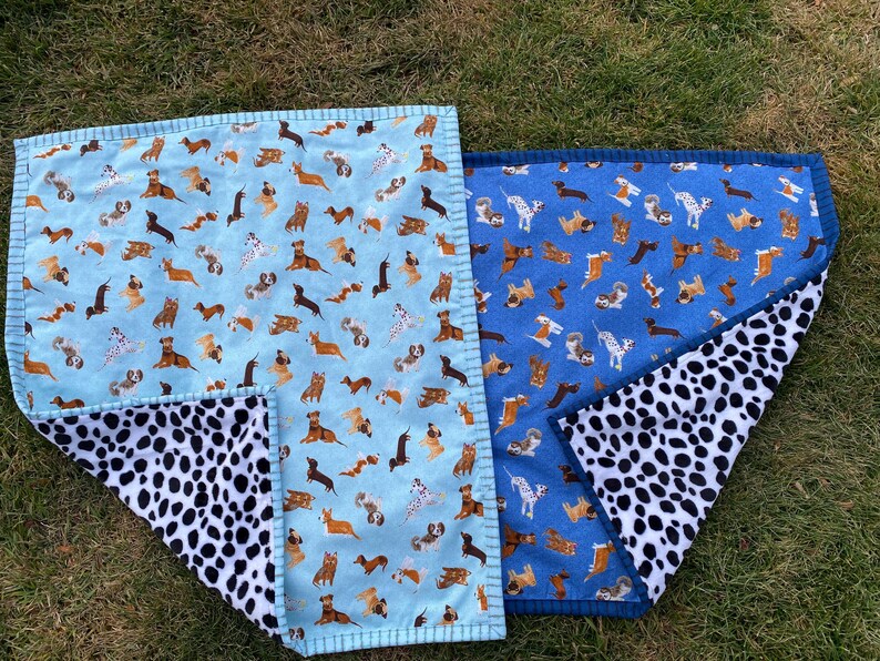 May include: Two dog-themed blankets. One is light blue with various dog breeds, the other is blue with similar dog illustrations. Both have a black and white dalmatian-spotted underside. The blankets are rectangular with a stitched border.