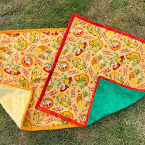 May include: Two Lion King themed baby blankets. The blankets feature a repeating pattern of Simba and Nala on a yellow background. One blanket has a yellow backing and orange trim, the other has a green backing and red trim.