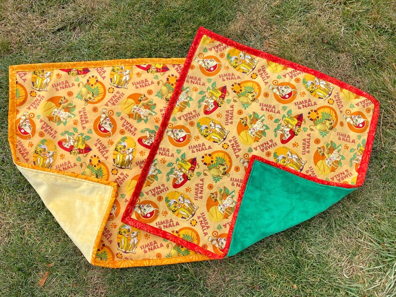 May include: Two Lion King themed baby blankets. The blankets feature a repeating pattern of Simba and Nala on a yellow background. One blanket has a yellow backing and orange trim, the other has a green backing and red trim.