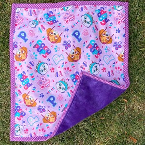 May include: A square, reversible blanket featuring a Paw Patrol theme. One side has a light purple background with Paw Patrol characters, paw prints, and the word "Paw Patrol". The other side is a solid, dark purple. The blanket is trimmed with a polka dot border.