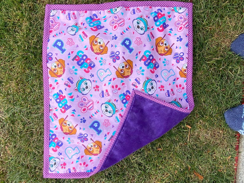 May include: A square, reversible blanket featuring a Paw Patrol theme. One side has a light purple background with Paw Patrol characters, paw prints, and the word "Paw Patrol". The other side is a solid, dark purple. The blanket is trimmed with a polka dot border.