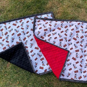 May include: A baby blanket featuring a pattern of Mickey Mouse on a white background with red and brown accents. The blanket has a black minky backing and a gray border. The blanket is laying on green grass.