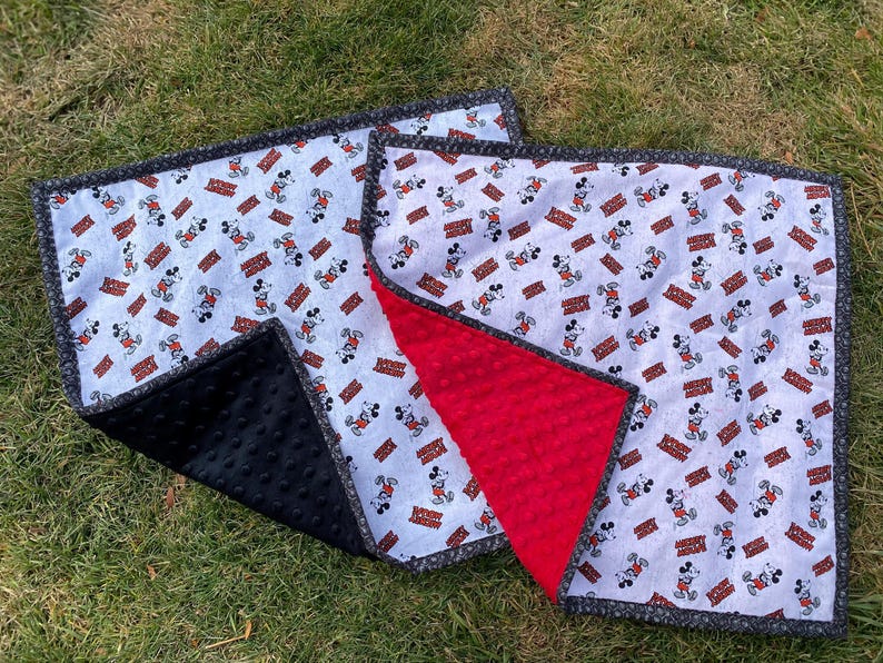 May include: A baby blanket featuring a pattern of Mickey Mouse on a white background with red and brown accents. The blanket has a black minky backing and a gray border. The blanket is laying on green grass.