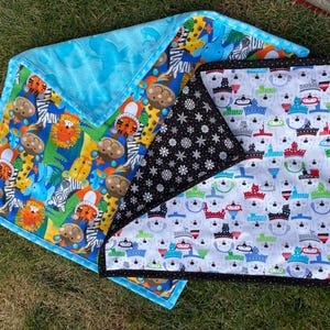 May include: A set of three baby blankets with colorful patterns. One blanket features a blue background with animal illustrations, another has a black background with white snowflakes, and the third has a white background with snowman illustrations. The blankets are on a green surface.