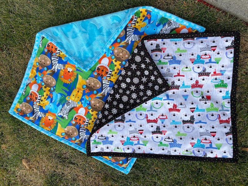 May include: A set of three baby blankets with colorful patterns. One blanket features a blue background with animal illustrations, another has a black background with white snowflakes, and the third has a white background with snowman illustrations. The blankets are on a green surface.