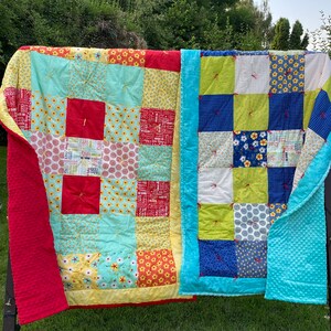 May include: Two colorful patchwork quilts hanging on a clothesline. The quilt on the left is red, yellow, blue, and green. The quilt on the right is blue, green, white, and yellow.