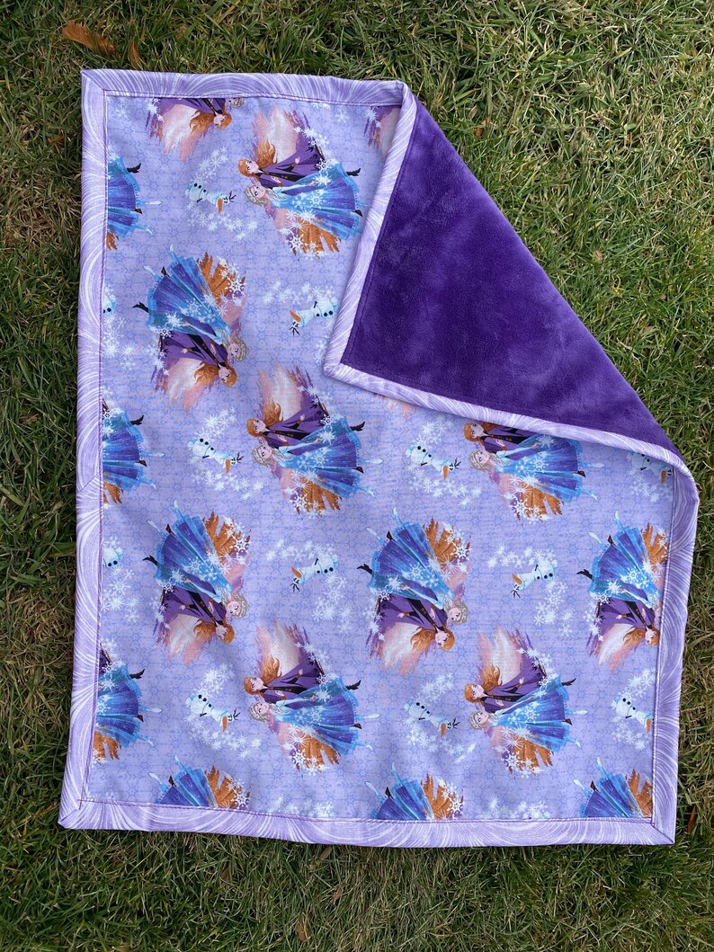 May include: A baby blanket featuring a purple and white pattern with images of a princess and a snowman. The blanket has a soft, plush purple backing and a patterned border.