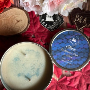 May include: A white candle with a blue glitter top in a silver tin. The candle is labeled "Neve Fir Balsam, Pine & Musk" and is 8 oz. of soy wax, hand poured.
