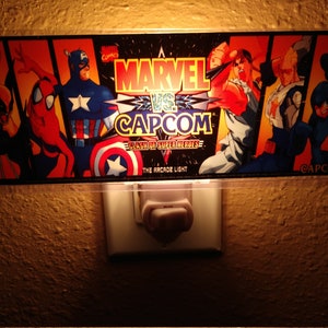 May include: A night light featuring a Marvel vs Capcom arcade game graphic. The graphic features characters like Captain America, Spider-Man, Wolverine, Ryu, and Mega Man. The text on the graphic reads "Marvel vs Capcom: Clash of Super Heroes - The Arcade Light".