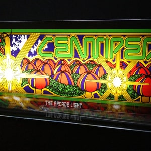Puede incluir: Un letrero de juego de arcade iluminado con un logotipo "CENTIPEDE" verde y amarillo, una serpiente verde y setas rojas y moradas. El letrero está iluminado con una luz amarilla brillante. El texto "THE ARCADE LIGHT" está en la parte inferior del letrero.