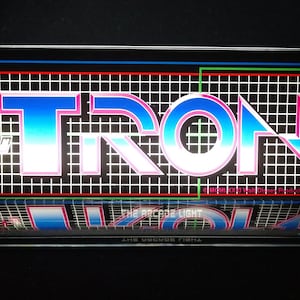 May include: A clear acrylic display case with a black background featuring the word "TRON" in a retro-style font with blue, white, and pink gradients. The word is surrounded by a grid pattern and a red, green, and blue oval. The text "THE ARCADE LIGHT" and "A MIDWAY MANUFACTURING CO. PRODUCT" are visible at the bottom of the display.
