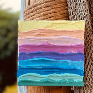 May include: A small, square canvas painting with a layered, abstract design. The painting features horizontal stripes of yellow, peach, pink, purple, blue, and teal paint. The paint is applied in a thick, textured style, creating a dimensional effect.
