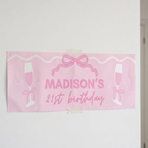 May include: A pink banner with the text "MADISON'S 21st birthday" in a cursive font. The banner features illustrations of champagne flutes, a bow, and a string of pearls. The banner is taped to a white wall.
