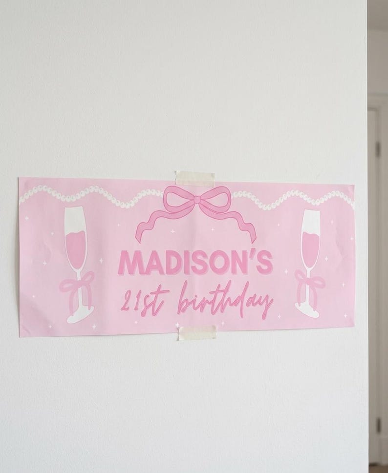 May include: A pink banner with the text "MADISON'S 21st birthday" in a cursive font. The banner features illustrations of champagne flutes, a bow, and a string of pearls. The banner is taped to a white wall.