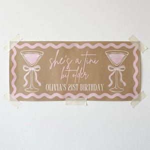 May include: A rectangular banner with a brown background and pink accents. The banner features two martini glasses with bows, a wavy pink border, and the text "she's a tini bit older" and "Olivia's 21st Birthday" in pink.