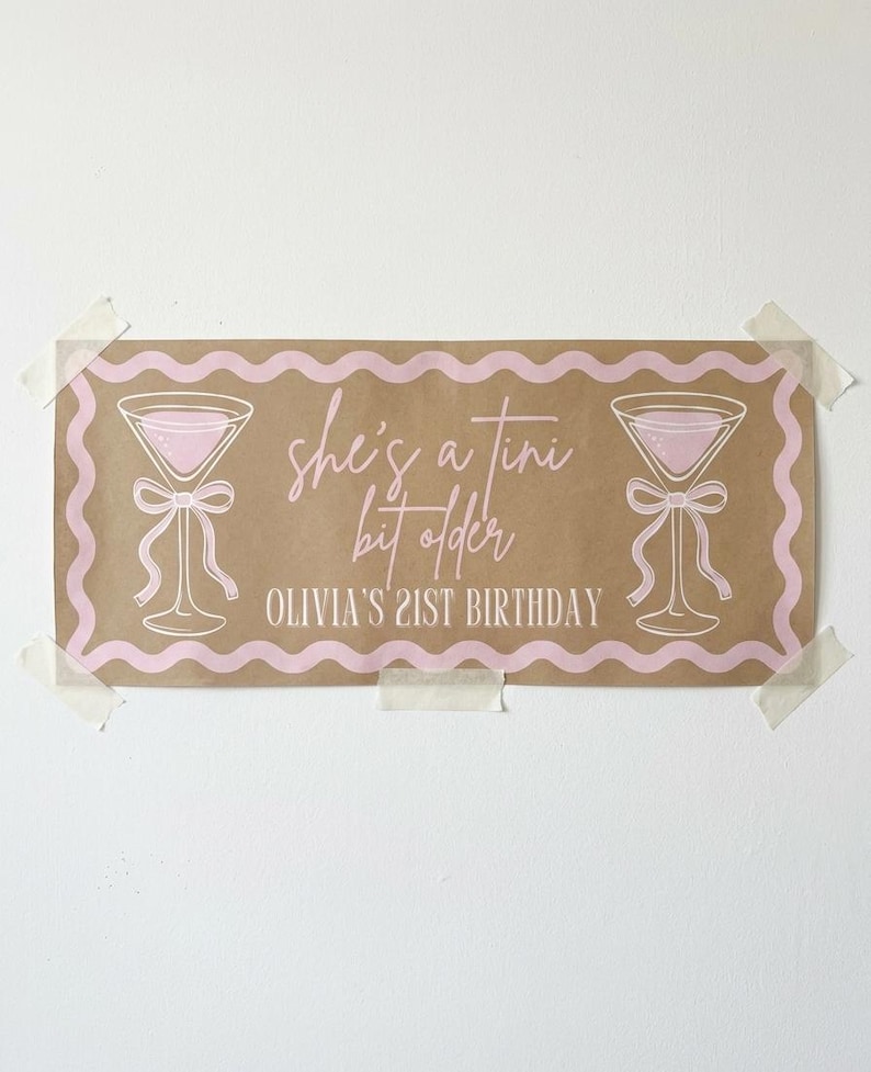 May include: A rectangular banner with a brown background and pink accents. The banner features two martini glasses with bows, a wavy pink border, and the text "she's a tini bit older" and "Olivia's 21st Birthday" in pink.