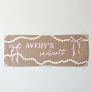 May include: A brown paper banner with the text "AVERY'S seventeenth" in pink script. The banner is decorated with pink bows and strings of white pearls. The banner is taped to a white wall.