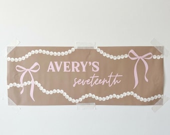 May include: A brown paper banner with the text "AVERY'S seventeenth" in pink script. The banner is decorated with pink bows and strings of white pearls. The banner is taped to a white wall.