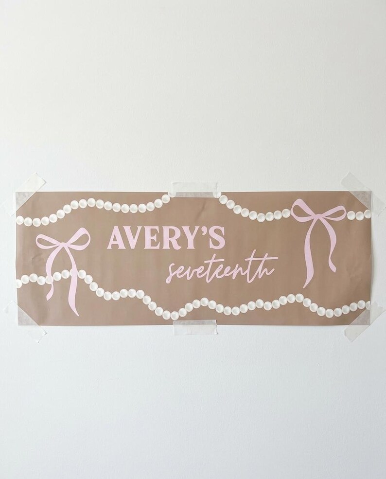 May include: A brown paper banner with the text "AVERY'S seventeenth" in pink script. The banner is decorated with pink bows and strings of white pearls. The banner is taped to a white wall.