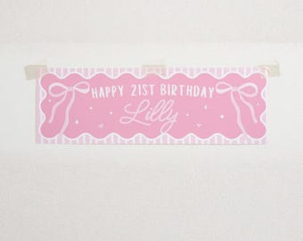 May include: A pink banner with the text "HAPPY 21ST BIRTHDAY Lilly" in white script. The banner has a scalloped edge and bow illustrations. It is taped to a white wall.